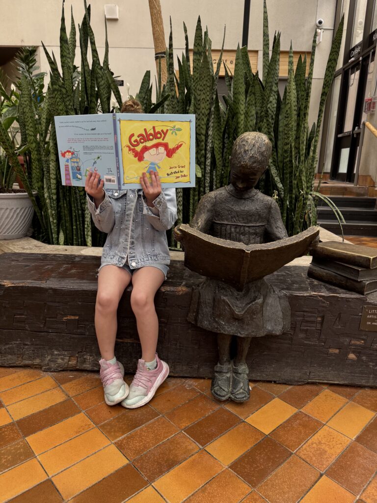 a girl reading gabby sits beside a bronze sculpture of a girl reading a book